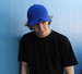 A blue cotton and spandex cap worn by a person in a black t-shirt, leaning against a blue wall.