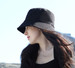 A woman wearing a black bucket hat with a wide brim, set against a bright outdoor background.
