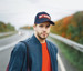 A navy cap with a logo features prominently on a man standing by a road, dressed in a navy jacket and red shirt.