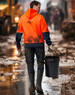A man wearing an orange and navy two-tone water-resistant fleece hoodie walks through muddy terrain, holding a bucket.