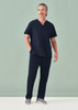 A man is wearing navy unisex scrub pants paired with a matching scrub top, standing against a light green background.