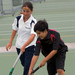 A white polo shirt with navy accents and a dark polo shirt with red accents, worn by two young athletes on a tennis court.