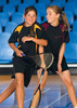Two girls smiling and playing tennis indoors, wearing black and yellow, and black and maroon Dri Gear T-shirts.