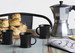 A silver espresso maker, three black mugs, and scones on a wooden table with a newspaper.