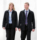 A black soft shell jacket worn by two individuals, one male and one female, against a light wooden background.