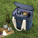 A drink cooler in navy blue with cream straps, containing various beverages and cups, set on grass beside snacks.