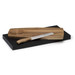 A wooden cheese board with a textured circular design, paired with a serrated knife, set on a black base.