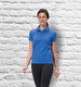 A woman in a blue ladies' polo shirt stands against a textured white wall, smiling. The shirt features a logo.
