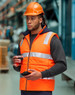 A man wearing an orange hi-vis hooded puffer vest and a hard hat stands in a warehouse, holding a coffee cup.