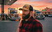 A man with a beard wears a tan and black trucker cap, standing beside trucks at a gas station during sunset.
