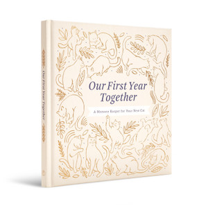 An angled, side view of the front cover of the fill-in, cat gift book, "Our First Year Together: A Memory Keeper for Your New Cat". The cover features the title in the center printed in blue foil, and is framed by a gold foiled cat design.