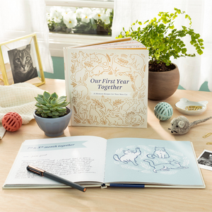 Lifestyle photo of the keepsake fill-in memory book of "Our First Year Together: A Memory Keeper for Your New Cat. The book is opened to a filled in page, sitting on a table with a picture of a cat and pet toys nearby.