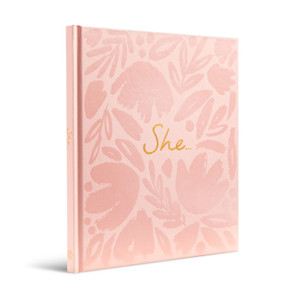 A side, 3/4 view of the front cover of inspiring friendship, women empowerment gift book, "She...", written by Kobi Yamada. The cover is pink with a floral pattern and the title "She..."{ embossed in gold foil.