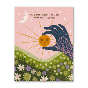 The front of this Birthday card features an illustration of a green hillside filled with flowers, a henna tattooed hand with fingers "pinching" a bright sun, against a light pink sky with stars and a moon, and reads "Each year makes you just more perfectly you." The card was illustrated by Lee Foster-Wilson.