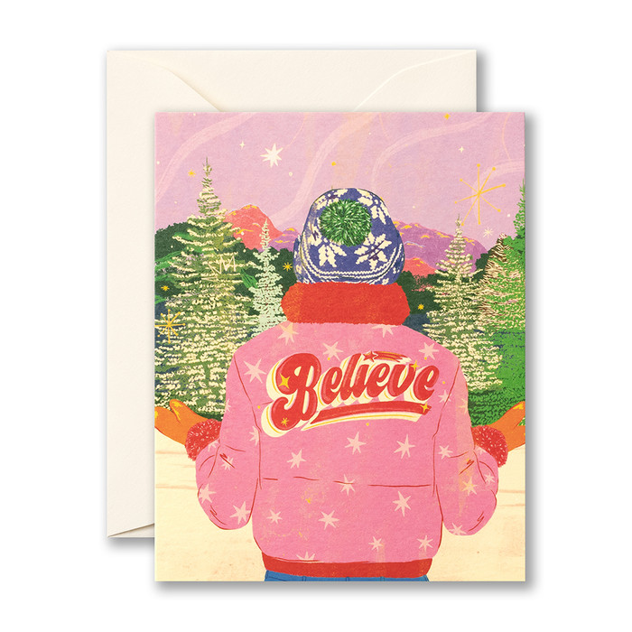 This image shows a holiday card with envelope. The card front features an illustration of a person standing among fir trees, wearing a wolly cap and bright pink jacket that "Believe" on the back.