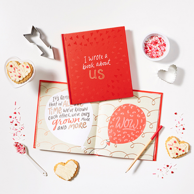 Lifestyle photo of the fill in gift book for your special sweetie, "I Wrote a Book About Us". The photo has the book opened to a page and has heart-shaped sugar cookies, cookie cutters, and confetti.