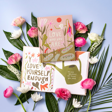 Lifestyle photo of the gift book "Love Who You Are", written by M.H. Clark and illustrated by Rafaela Pascotto. Book is open to a page with the encouraging, uplifting statement: "Love yourself enough to give yourself the things you need." The book is placed around an assortment of fresh flowers.