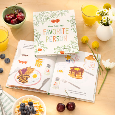 This lifestyle image shows the love & friendship gift book "You Are My Favorite Person". The scene is at a breakfast table with a bowl of cherries, waffles, blueberries, orange juice, fresh flowers, and the book is open to a page illustrated with breakfast treats.