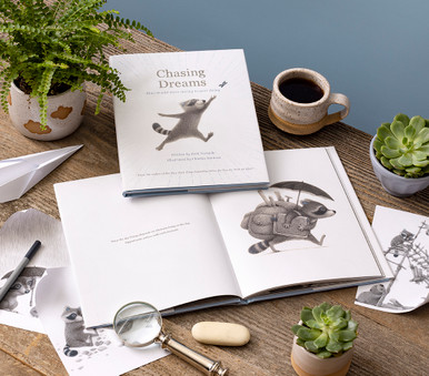 This image shows the front view of the of the gift book, Chasing Dreams: How to add more daring to your doing, written by Kobi Yamada and an inside spread from the book of the adorable raccoon character rushing off with a filled backpack, umbrella and book. The copy on the page reads "What the day brings depends on what you bring to the day. Opportunity arrives with each moment."