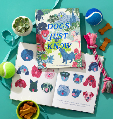 A lifestyle image that has dog treats, toys and accessories placed around the gift book "Dogs Just Know". A bright and cheerful cover with an illustratation of five happy dogs in a setting with flowers and green bushes. This book is written by M.H. Clark and illustrated by Emily Taylor.