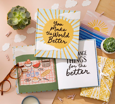 A lifestyle image of the gift book "You Make the World Better". The book cover is featured and a open to a page that shows an illustrated picnic scene.