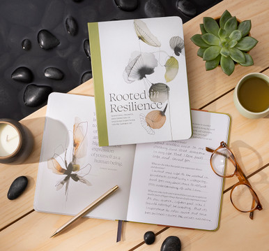 This image shows the front view of the activity book "Rooted in Resilience" - A Guided Journal of Questions, Prompts, and Exercises to Discover Everything You’re Capable of.  The notebook is sitting on a light wood table with succulents, candles and a soothing water feature surrounding the book.