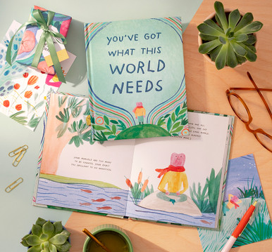 This lifestyle image shows the front view of  "You've Got What This World Needs" and an inside spread from the book. Surrounding the book is a wrapped gift and succulent plants.