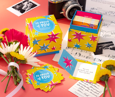 This image shows a lifestyle photo featuring "So Proud of You" ThoughtFulls for kids. The scene includes flowers, a sheet of music, recital photos, and a camera surrounding a box and open cards of ThoughtFulls pop open cards for kids.