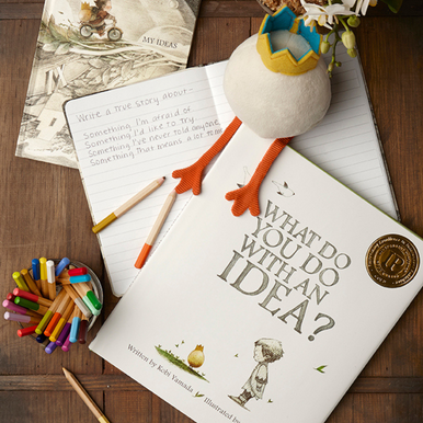 Lifestyle photo featuring the all-ages picture book, What Do You Do With an Idea, written by Kobi Yamada and illustrated by Mae Besom, and the companion idea plush stuffy.