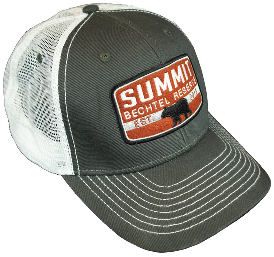 Trucker Cap with white mesh back and dark gray front.  A rust orange colored patch front center that says, Summit Bechtel Reserve, has a black bear in the center of the patch with the establish date of 2013 and has a plastic snap closure. -Side View