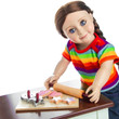 Against white background an 18 inch doll leans over the cookie baking set on the doll kitchen table.
