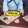 In 18 inch doll kitchen on farmhouse table, a doll holds the rolling pin over a wooden breadboard, with the 2 cookie cutters and 4 star and heart shaped sugar cookies.