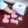 A doll's hand reaches toward a selection of toy cookies arranged on a dark red surface next to a white bakery box. The assortment includes three heart-shaped cookies textured with pink sprinkles and three star-shaped cookies iced in light purple. The white rectangular box behind them features a prominent decorative crest logo printed on its lid.