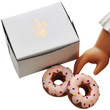 On white background there are 2 doll sized pink doughnuts and a bakery box with the Queen's Treasures fleur-de-lis embossed on top. A doll hand is seen reaching in for a doughnut.