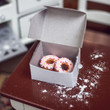 In 18 inch doll kitchen there are two pink frosted doughnuts inside white bakery box on kitchen table.