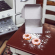 In 18 inch doll kitchen there are two pink frosted doughnuts with opened white bakery box on a kitchen table.