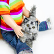 A doll wearing a bright horizontally striped t-shirt and blue jeans sits on a plain white surface, holding a realistic fluffy grey-and-black striped toy cat. The toy cat has bright green eyes and a raised tail, sitting between the doll's legs.