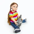 A doll with long brown hair and blue eyes sits on a plain white background, holding a realistic fluffy gray-and-black striped toy cat between her legs. The doll is dressed in a bright horizontally striped rainbow t-shirt, dark blue jeans, and sneakers with black soles, while the small toy cat features a raised tail.