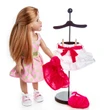 An 18-inch doll with long red hair stands against a plain white background, looking towards a dark wooden clothing display stand. The doll wears a sleeveless pink dress with green polka dots and matching pink sneakers. A white tiered skirt with a hot pink bow is clipped onto the stand, while a matching hot pink top hangs off to the side, and a hot pink purse rests on the round base.