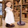 An 18-inch doll with brown hair styled in two long braids stands on a dark wooden display base against a solid white background. She is turned slightly to the side, wearing a long-sleeved, vintage-style white nightgown with a ruffled collar and cuffs. There is an empty doll stand. Both are in a store from the 1900's
