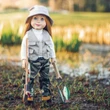 An 18-inch doll with blue eyes and straight red hair stands outdoors in a marshy, grassy field dressed in a complete fishing outfit. She wears a tan bucket hat, a matching grey multi-pocket utility vest over a long-sleeved white shirt, green camouflage waders, and boots. In one hand, she holds a miniature wooden fishing rod, and in the other, she holds a small mesh fishing net containing a tiny painted toy fish against a softly blurred sunlit background.