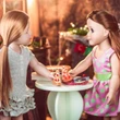Two 18-inch dolls stand facing each other at a small, light-colored pedestal table, sharing a slice of toy cheese pizza. The doll on the left has long, straight blonde hair and wears a textured light grey ruffled dress, while the doll on the right has long brown hair and wears a pink dress with green polka dots. In the background, a warm, cozy room is softly blurred with glowing fairy lights decoration.
