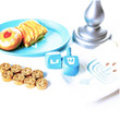 A close-up shot of a miniature Hanukkah play food and accessory set is displayed on a bright white surface. In the upper left, a teal plate holds a realistic toy jelly donut and a golden-brown latke. To the right, the metallic silver base of a play menorah sits next to a folded white fabric bag printed with a teal menorah logo. In the center, two light blue wooden dreidels rest beside a neat double row of eight tiny, textured gold play coins.