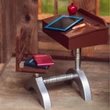 A side view captures a miniature vintage-style school desk with an attached chair, set on a light wood floor inside a rustic dollhouse room. A toy slate board with a blue border, a small wooden pencil, and a red apple model are arranged on the slanted brown desktop. A small red notebook and a blue book are stacked on the wooden seat of the chair, which connects to the desk via a metallic silver painted base frame.