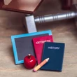 A close-up shot shows a collection of miniature school accessories resting on a light wood floor beneath a toy desk frame. The set includes a small black slate with a blue border, a red "ARITHMETIC" book, and a dark blue "HISTORY" book stacked together. A small red toy apple and a short yellow pencil lie on the floor in front of the books.