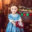 An 18-inch doll with long brown hair stands facing forward in a cozy holiday room, holding a miniature red gift box decorated with white snowflakes and a white ribbon. She is wearing a blue-and-white patterned dress featuring a white lace collar and cuffs. The background shows a warmly lit fireplace decorated with a green and red banner and soft, glowing Christmas lights.
