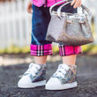 An 18-inch doll wears silver glitter high-top sneakers, dark denim jeans with pink plaid cuffed hems, and holds a matching silver glitter handbag. The lower half of the doll is positioned on a paved surface against a blurred background featuring a white fence.