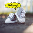 A pair of miniature silver glitter high-top sneakers with white laces and toe caps rests on a dark pavement. One shoe stands upright while the other leans slightly against it. Above them, a bright yellow comic book-style speech bubble contains the hand-written word "Retiring!". The background shows a soft, out-of-focus outdoor setting with white fencing.