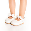 A close-up view shows a doll's feet wearing a pair of clean white Mary Jane-style dress shoes against a solid white background. The shoes feature delicate ankle straps adorned with prominent white bows and have thin, light brown outsoles.