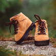A pair of miniature tan suede doll hiking boots sits outdoors on a textured stone surface with a blurred green background. The boot on the left leans at a slight angle against the right boot, showcasing dark brown laces threaded through silver metal eyelets and thick, black rugged rubber soles with heavy treading.